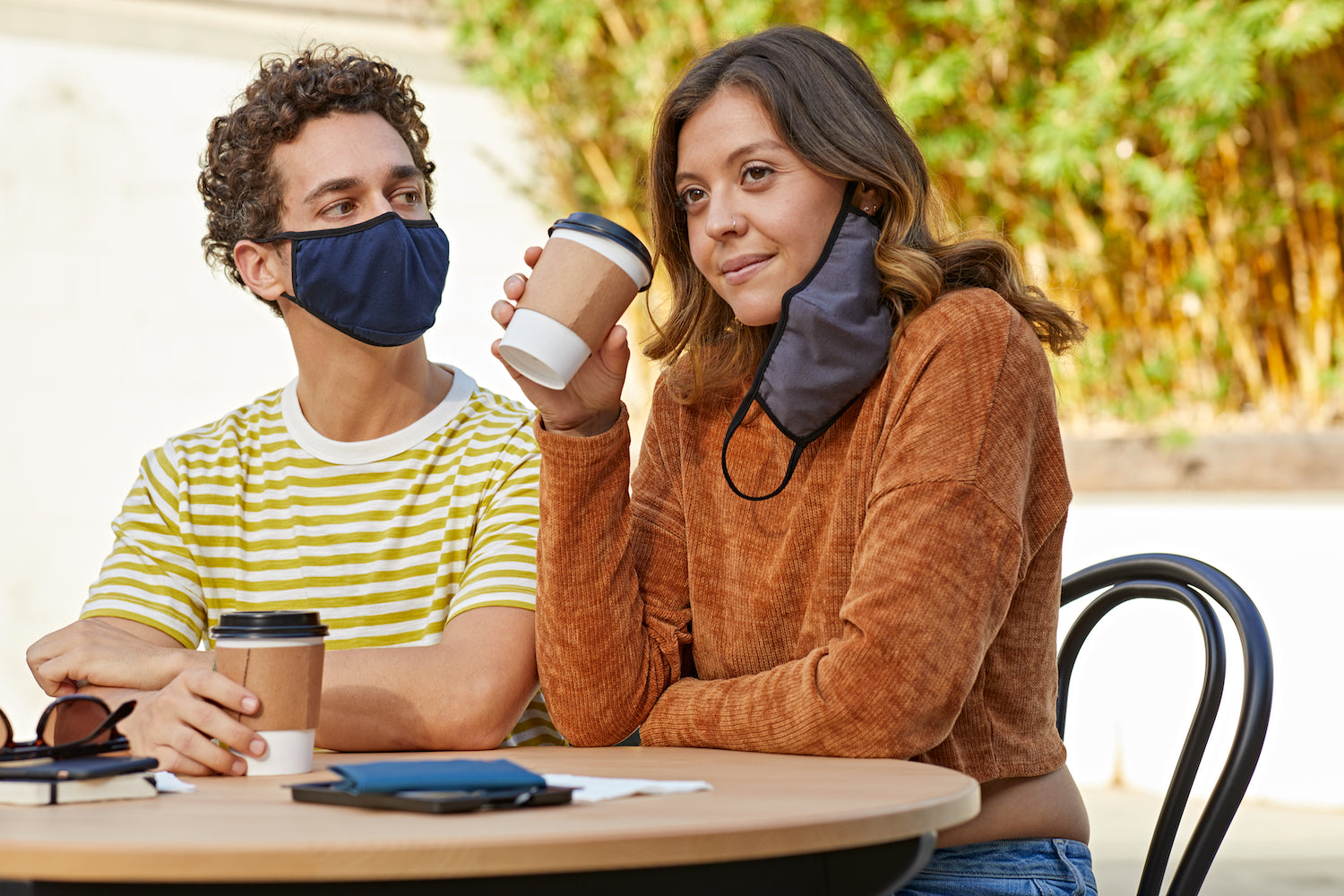 Man and woman outdoors. Man is wearing a cloth mask and looking at the woman, who has taken her mask off and it is hanging by her ear as she drinks a coffee.