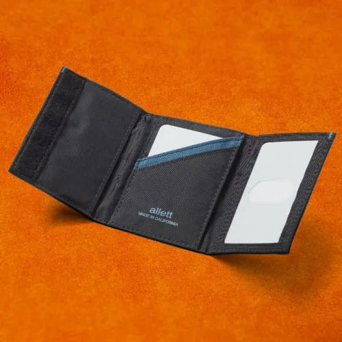 Picture shows a slim black men's trifold wallet with velcro open on an orange background. Wallet is made from Allett in California.