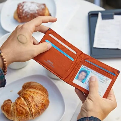 Person with a tattoo on their left hand opens an ID holder wallet that displays an ID through a clear case. Wallet is from Allett Wallets.