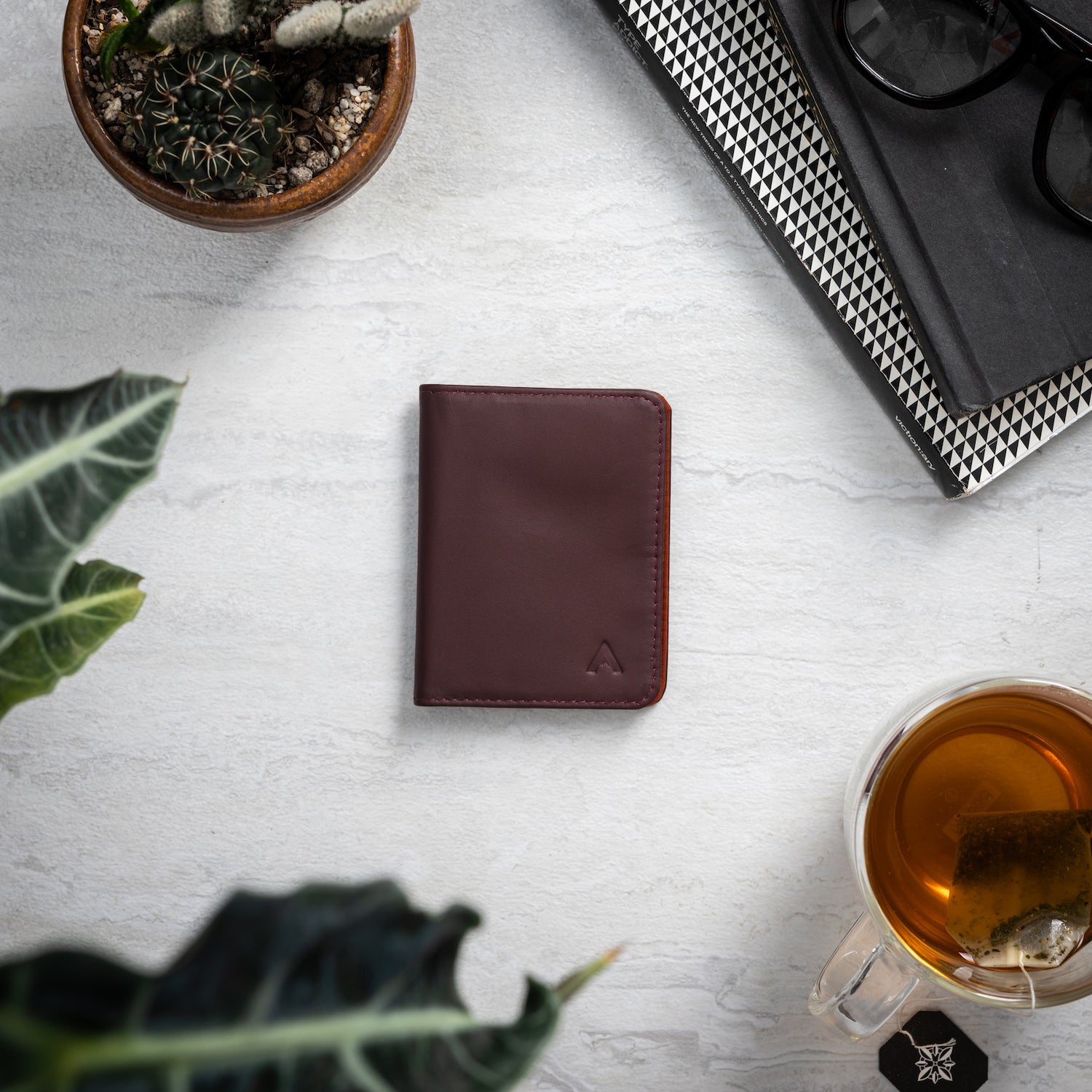 Closed merlot hybrid card wallet on a white background surrounded by items such as a glass tea cup, notebooks, and cactus. Allett hybrid card wallet leather merlot