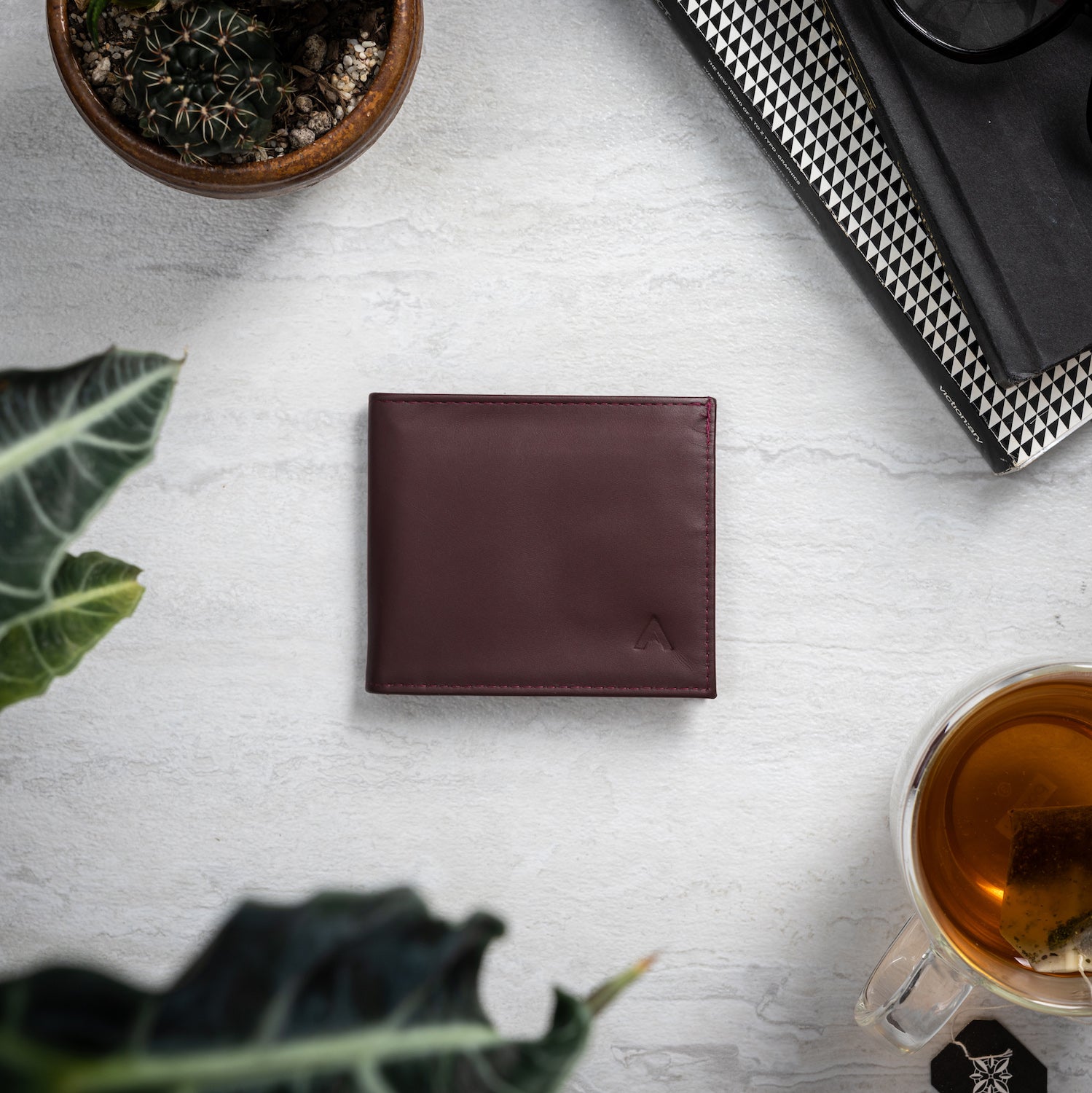 Allett's merlot colored Nappa ultra thin leather wallet on a light grey table, closed, surrounded by household items like greenery and a coffee mug. Allett id wallet leather merlot