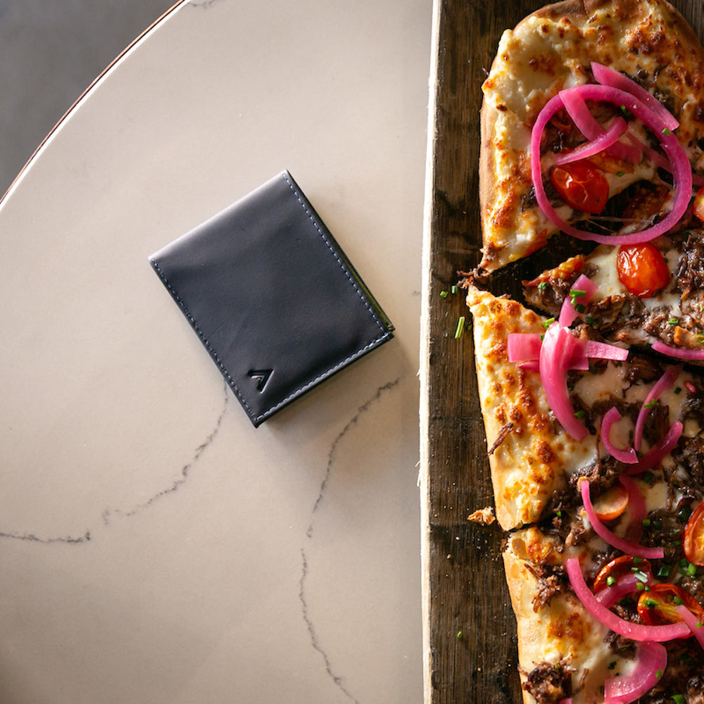 Allett's premium ultra slim leather sports wallet next to a flatbread pizza on a table. Allett Leather Sport Wallet- midnight-blue