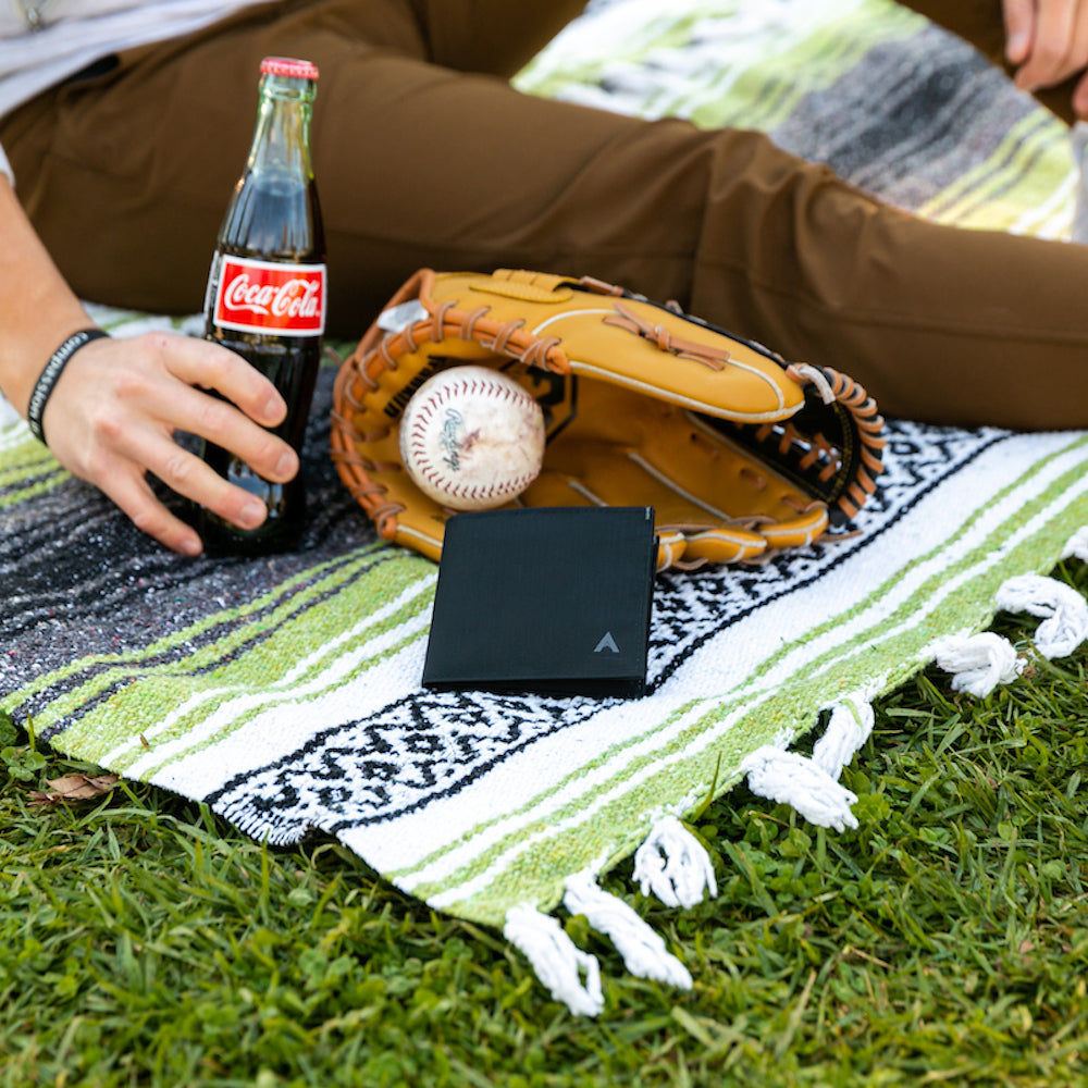 Crop of a person's hand outdoors holding a coke bottle with their wallet on a mat on the grass next to a baseball glove and baseball - Allett ID Wallet Nylon RFID jet-black 